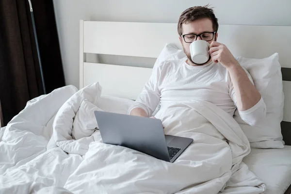 Seriös man läser nyheterna på laptop och dricker kaffe, sitter i sängen, på morgonen, i naturligt ljus. Modern livsstil. — Stockfoto