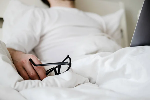 Hand håller i glasögon på en mjuk, vit filt. Mannen arbetade hela natten med en laptop i sängen och somnade av trötthet på morgonen. — Stockfoto