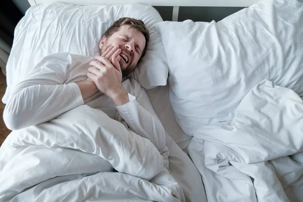 Mannen i vit pyjamas lider av svår tandvärk, han håller handen på kinden, liggande i sängen, i huset, i naturligt ljus. — Stockfoto
