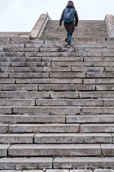 Turista en ropa de abrigo y con una mochila urbana sube los antiguos escalones de piedra de una escalera alta en un día de invierno. Vista inferior . — Foto de Stock