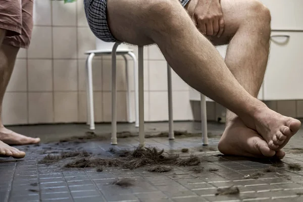 El hombre con las piernas cruzadas se sienta en una silla, su pelo recortado está acostado junto a él, en el suelo de baldosas en el baño . — Foto de Stock