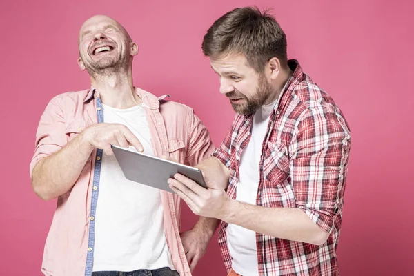 Man laughs while watching a video on a tablet, his friend looks in shock at the screen and smiles. Positive emotions. — Stock Photo, Image