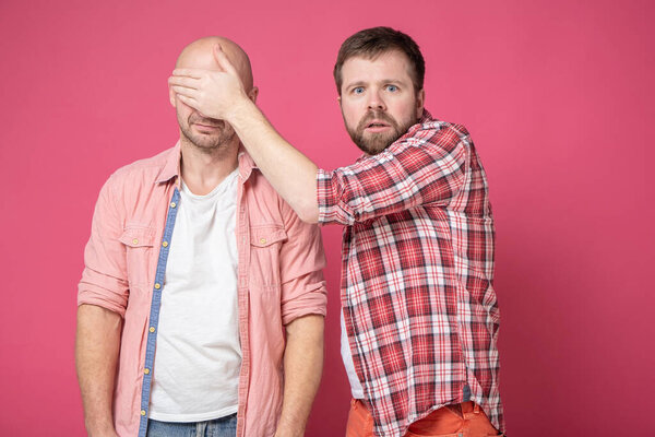 Excited bearded man holds hand in over of his friend eyes and looks frightened at the camera.