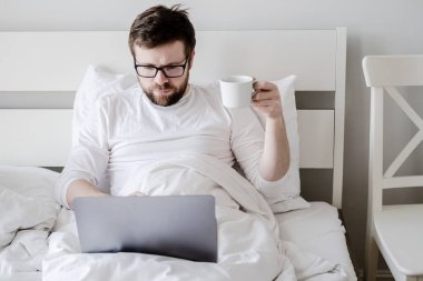 Focused bearded man, with a cup of coffee in his hand, works on a laptop, sitting in bed, under natural light. Modern lifesty.