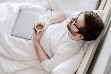 Relaxed man with a cup of coffee in hands, sitting in bed, next to a closed laptop. Top view.