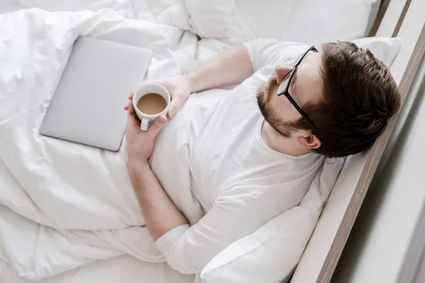 Avslappnad man med en kopp kaffe i händerna, sitter i sängen, bredvid en stängd laptop. Ovanifrån. — Stockfoto