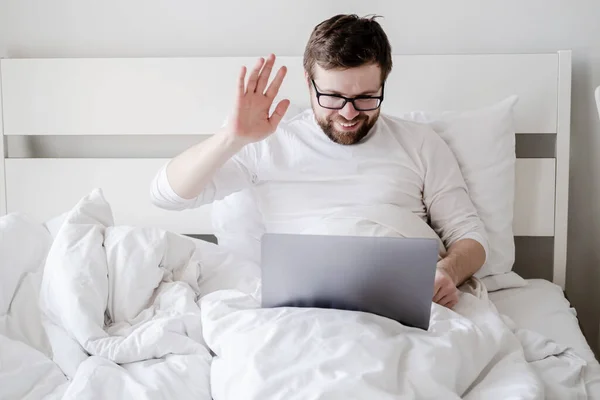 Människan kommunicerar på distans genom ett videosamtal från en bärbar dator, hon tittar på skärmen, ler och vinkar hand medan hon sitter i sängen. — Stockfoto