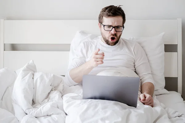 Missnöjd, arg man kommunicerar på distans via ett videosamtal från en bärbar dator, sittandes i sängen. — Stockfoto