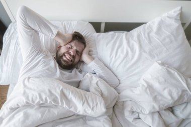 Man in bed suffers from a headache or a hangover, he clenches head in his hands and grins his teeth from pain.