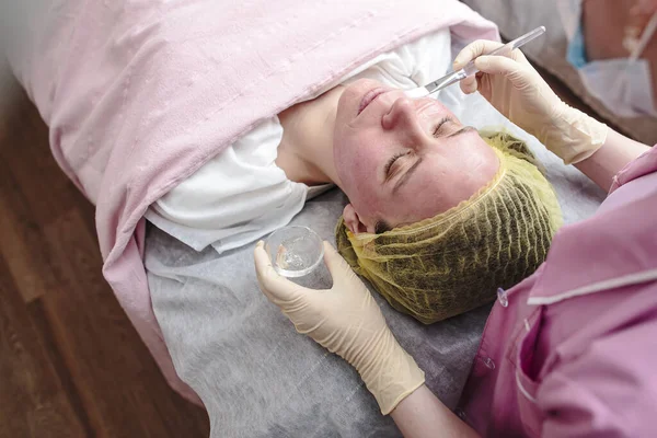 Kosmetolog nakłada kojącą, przezroczystą maskę na zaczerwienioną skórę twarzy pacjenta, po peelingu chemicznym. — Zdjęcie stockowe