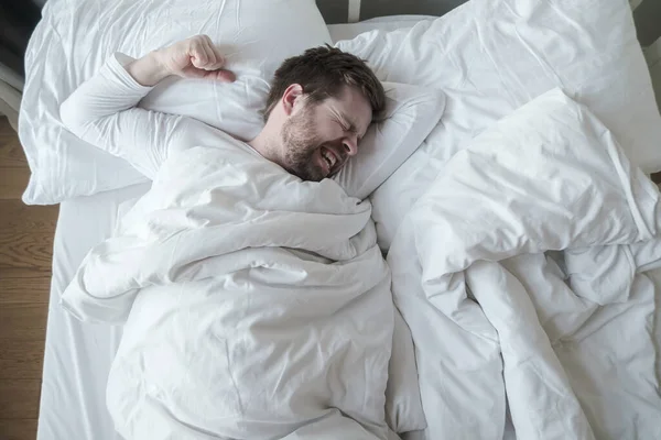 El hombre en pijama blanco tiene una pesadilla terrible, rechina los dientes y prepara un puño para la protección, acostado en su cama . — Foto de Stock