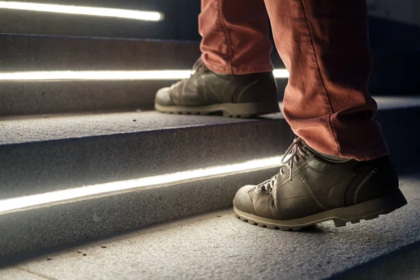 Male legs in walking boots climb the lit stone steps on a winter evening.