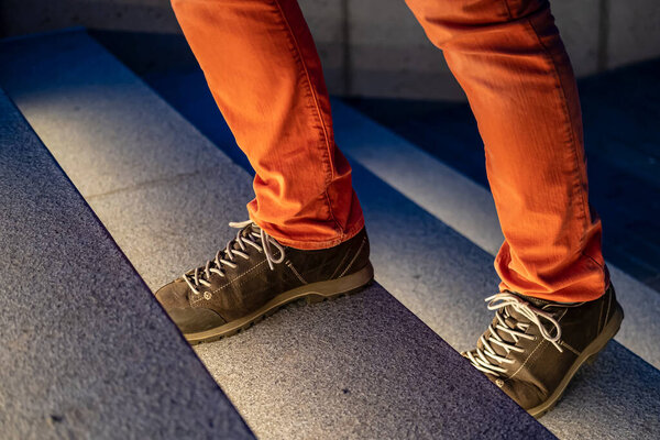 Male legs in walking boots and red jeans climb the illuminated stone steps on a winter evening. Close-up.