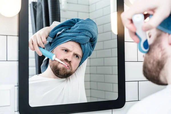 Sleepy man with a towel on head brushes teeth with an electric brush and looks with indifference at his reflection in the mirror. — Stock fotografie