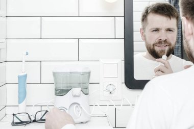 Cute man cleans his teeth with the help of electric oral irrigator with a jet of water, looking in the mirror. Copy space.