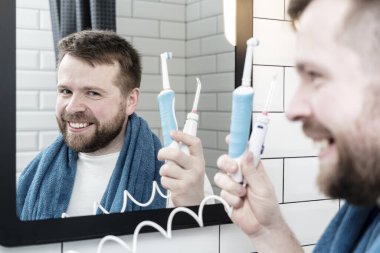 Positive man holds a rechargeable toothbrush in hand and an electric, oral irrigator, looks in the mirror and smiles.