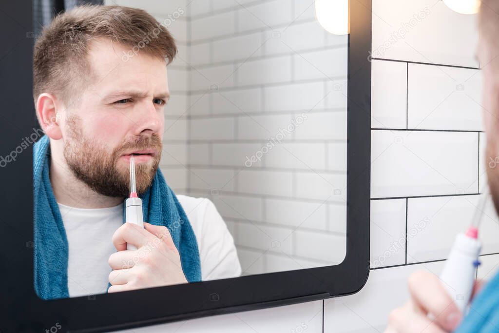 Serious man brushing his teeth with an electric oral irrigator with a jet of water, looking in the mirror.