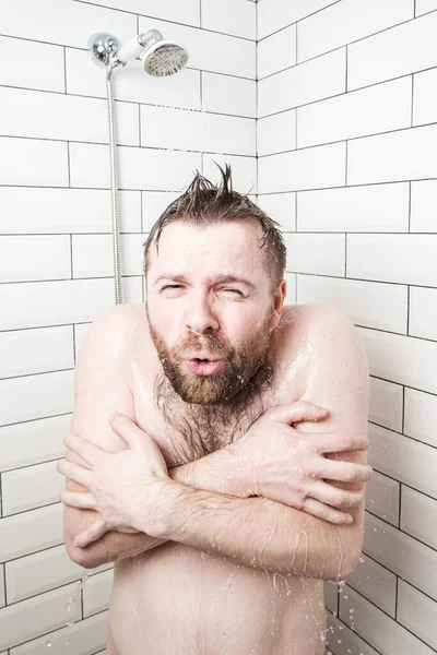 Man, with a funny expression on his face, feels shocked at taking a cold shower, he froze and tries to cover his body with hands. — Stock fotografie