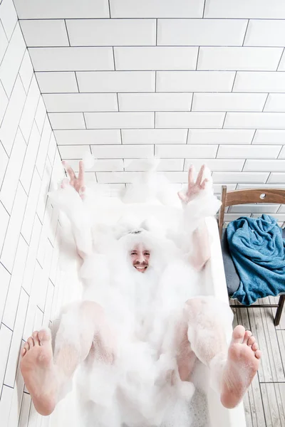 Piacevole, divertente uomo caucasico si trova in una vasca da bagno con schiuma lussureggiante, sorride e agita le braccia e le gambe. Concetto di pulizia . — Foto Stock