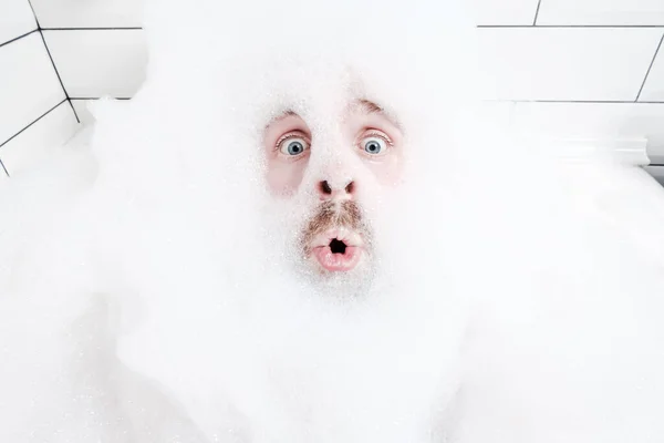 Rosto masculino surpreso em uma espuma exuberante, apenas olhos e boca são visíveis, close-up. O homem gosta de se deitar na banheira, com água ensaboada . — Fotografia de Stock