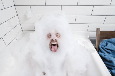 Funny, positive man lies in a bathtub with lush foam and shows his tongue.