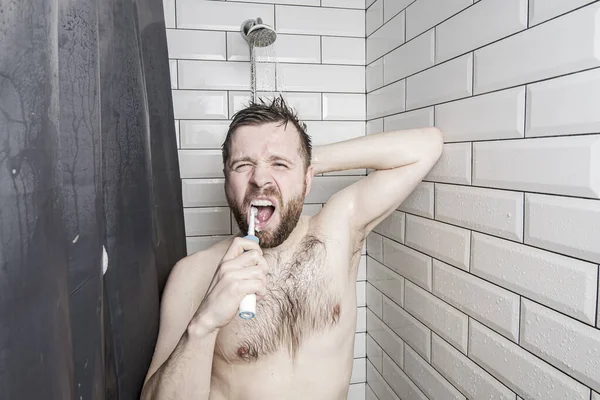 El hombre complaciente cepilla los dientes con un cepillo de dientes recargable mientras está de pie en la ducha, bajo el agua corriente, en el baño . — Foto de Stock