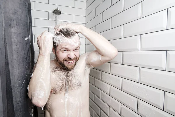 Hombre satisfecho, enjabonado con espuma, se lava en la ducha, bajo un chorro de agua, sonríe y mira a la cámara . — Foto de Stock