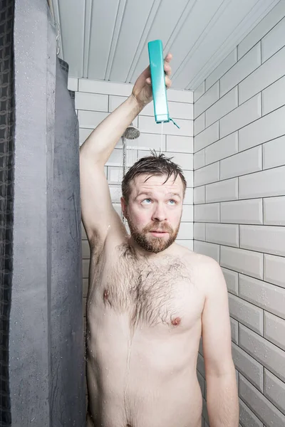 Hombre enfocado derrama champú en la cabeza, de pie en la ducha, bajo un chorro de agua, contra una pared de azulejos blancos . — Foto de Stock