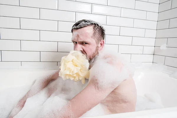 Displeased, weird man with a soapy sponge puff in mouth, sits in a bathtub with lush foam and looks angrily at the camera.