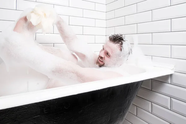 Soddisfatto, uomo sorridente in una schiuma saponata lussureggiante, lava il piede con entusiasmo con l'aiuto di un soffio di spugna, seduto nel bagno — Foto Stock