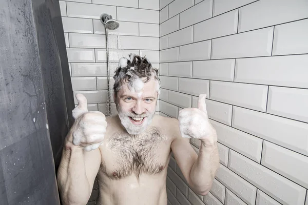 Satisfied man, soaped with foam, washes in the shower, under the stream of water, smiles and shows a thumb up gesture.