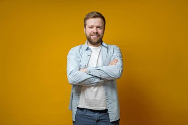 Kendine güvenen sakallı adam kollarını kavuşturmuş, tatlı tatlı gülümsüyor ve gözlerini kıstırarak kameraya bakıyor..