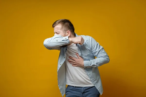 L'uomo con una maschera medica tossisce al gomito e tiene la mano sul petto, provando dolore. Concetto di diffusione del virus . — Foto Stock