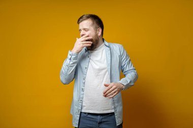 Sleepy bearded man yawns, covering mouth with hand and closing eyes.
