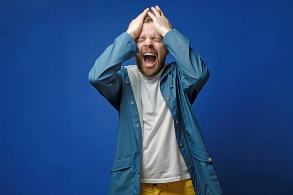 Handsome man is very upset, he holds hands on head and screams loudly, on a blue background. Mental health concept. — Stockfoto