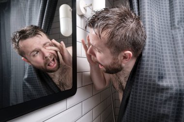 Wet man experiences discomfort, he looks in the mirror and tries to get an eyelash that got into the eye, standing in shower.