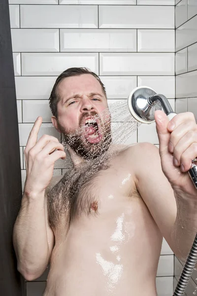 Homem barbudo engraçado canta no banheiro, usando uma cabeça de chuveiro com água corrente em vez de um microfone. Estilo de vida . — Fotografia de Stock