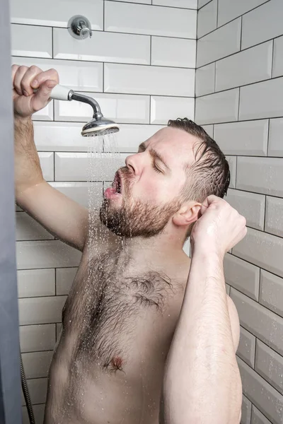Un hombre divertido canta con entusiasmo en el baño, usando una ducha con agua corriente en lugar de un micrófono. Estilo de vida . — Foto de Stock
