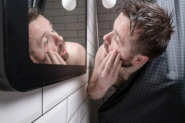 Wet man looks in the mirror his eyes, reddened by fatigue, standing in the shower, behind the curtain. — Stockfoto