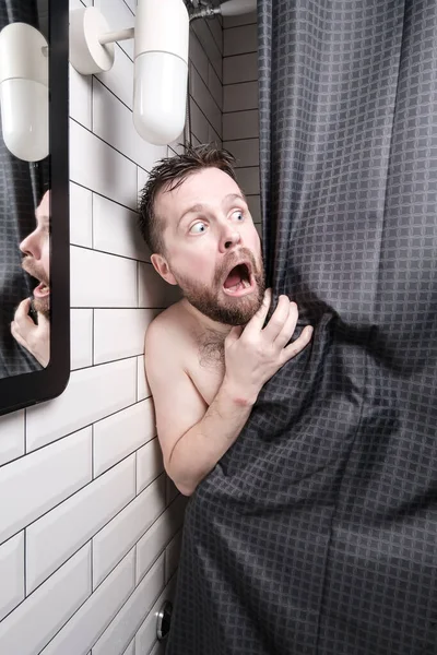 Frightened man peeks out from behind the curtains, in the shower, he is shocked, opened mouth and makes big eyes.