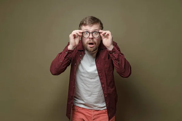 Surprised bearded man, in a burgundy shirt, in amazement holds glasses in hands and looks with big eyes into the camera. — Stock Photo, Image