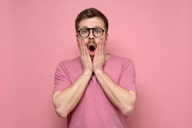 Komik gözlüklü yakışıklı adam şaşırır, elleriyle yüzünü tutar, ağzı açık bir şekilde kameraya bakar..