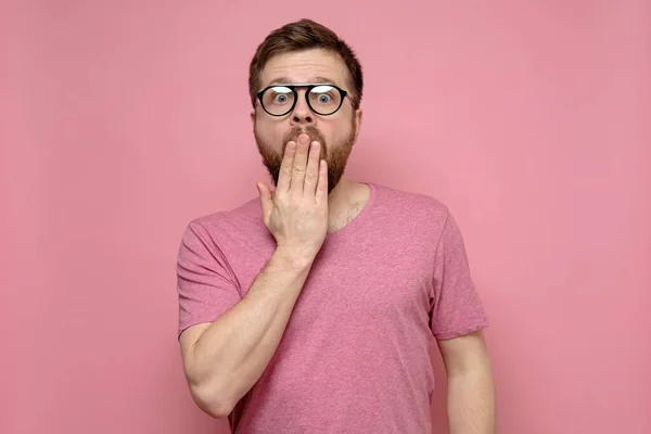Hombre barbudo con gafas está asustado y sorprendido, cubrió la boca con la mano y mira a la cámara, sobre un fondo rosa . — Foto de Stock