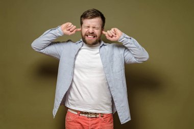 Gürültülü bir sesten rahatsız olan adam, kulaklarını işaret parmaklarıyla kapatıyor, dişlerini hoşnutsuzlukla sırıtıyor, yeşil bir arka planda..