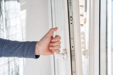 Hand opens a PVC window, holding the handle in an upright position, with a fixing device attached to the frame for ventilation.