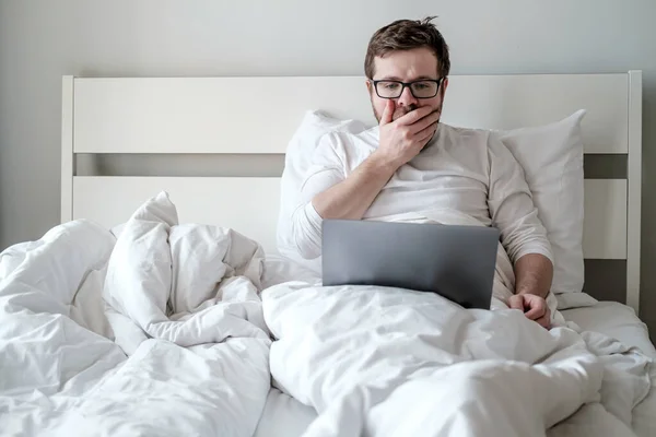 Mannen med en laptop i sängen fick dåliga nyheter, han är rädd och upprörd. Begreppet distansarbete under karantän. — Stockfoto