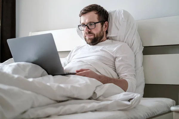 Mannen i pyjamas arbetar med laptopen på distans medan han sitter i sängen. Begreppet karantän under en virusepidemi och en pandemi. — Stockfoto