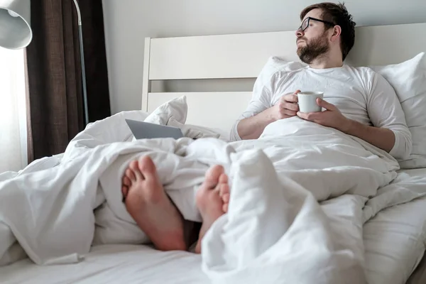 Pensive man med en kopp kaffe i händerna vilar, han arbetar på distans med en bärbar dator, sitter i sängen. Begreppet karantän. — Stockfoto