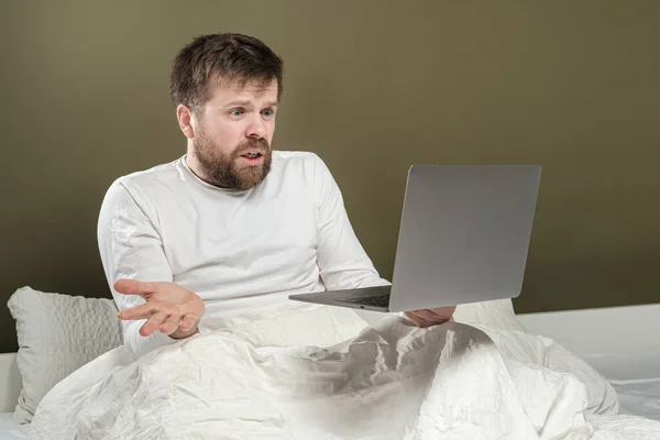 Människan arbetar med en bärbar dator på distans medan hon sitter i sängen och pratar känslomässigt med anställda via videosamtal. Karantänkoncept. — Stockfoto