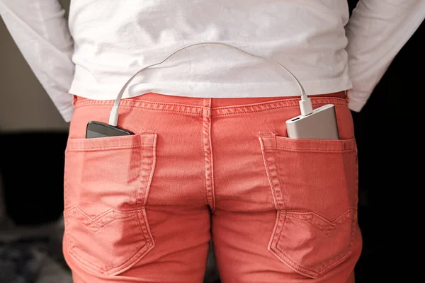 Power Bank liegt in der Gesäßtasche einer korallenfarbenen Jeans, und in einer anderen befindet sich ein Handy, das geladen wird. — Stockfoto